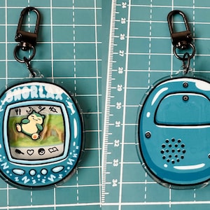 May include: A blue and white Snorlax Tamagotchi keychain with a black metal clasp. The front of the keychain shows Snorlax sleeping on a green background. The back of the keychain is solid blue with a speaker.