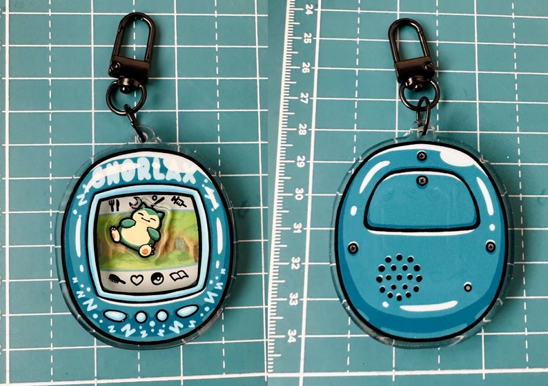 May include: A blue and white Snorlax Tamagotchi keychain with a black metal clasp. The front of the keychain shows Snorlax sleeping on a green background. The back of the keychain is solid blue with a speaker.