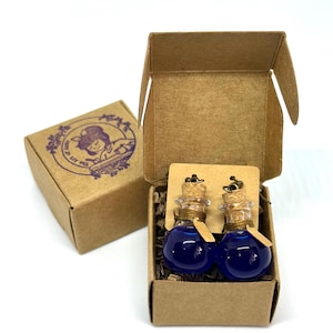 May include: Two small glass potion bottle earrings with cork stoppers and blue liquid, presented in a small open cardboard box. A second closed box with a purple illustration is visible. The earrings are displayed on a brown card.