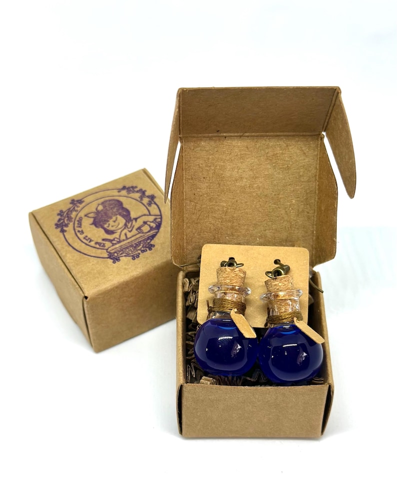 May include: Two small glass potion bottle earrings with cork stoppers and blue liquid, presented in a small open cardboard box. A second closed box with a purple illustration is visible. The earrings are displayed on a brown card.