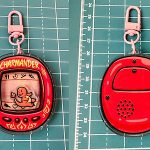 May include: A red and clear plastic keychain featuring a Charmander character from the Pokemon franchise. The keychain has a small screen with a cartoon image of Charmander and the text "CHARMANDER" on the front. The back of the keychain is solid red with a black speaker.