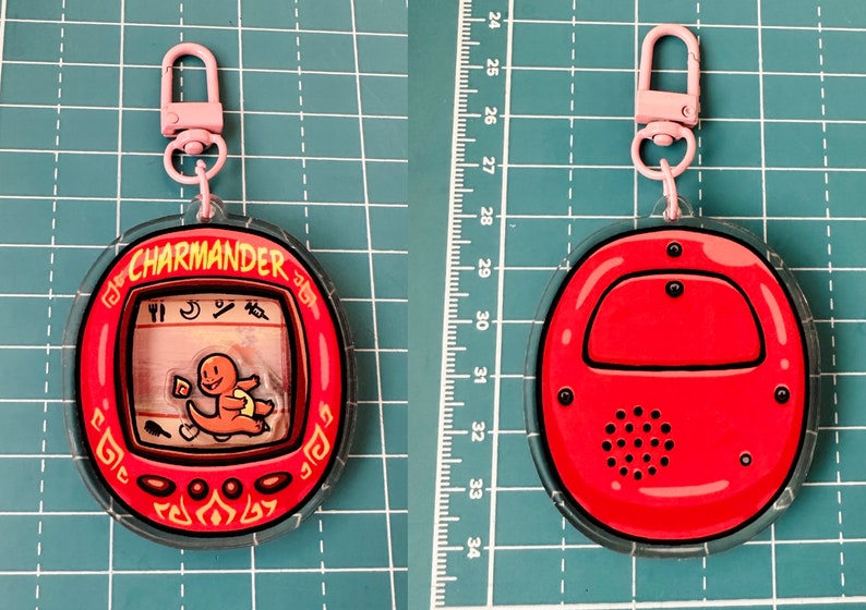 May include: A red and clear plastic keychain featuring a Charmander character from the Pokemon franchise. The keychain has a small screen with a cartoon image of Charmander and the text "CHARMANDER" on the front. The back of the keychain is solid red with a black speaker.