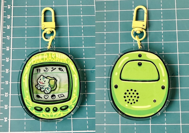 May include: A green and yellow Pokemon-themed keychain with a clear plastic casing. The front of the keychain features a digital pet screen with the text "BULBASAUR" and an image of the Pokemon character. The back of the keychain is plain green.