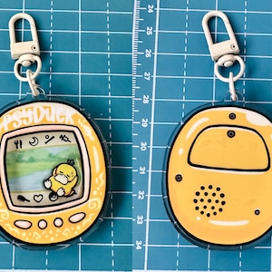 May include: A yellow and white plastic keychain shaped like a Tamagotchi with a cartoon Psyduck on the screen. The keychain has a white metal clasp.
