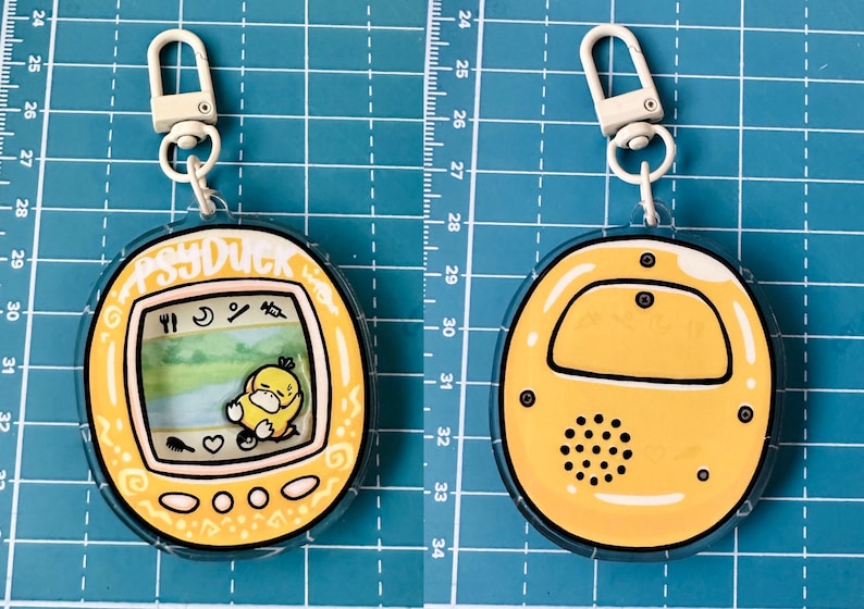 May include: A yellow and white plastic keychain shaped like a Tamagotchi with a cartoon Psyduck on the screen. The keychain has a white metal clasp.