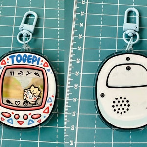 May include: A white and blue plastic keychain with a Togepi character from the Pokemon franchise. The keychain is shaped like a Tamagotchi and has a small screen with a cartoon image of Togepi. The keychain has a blue clip and a black and white speaker on the back.
