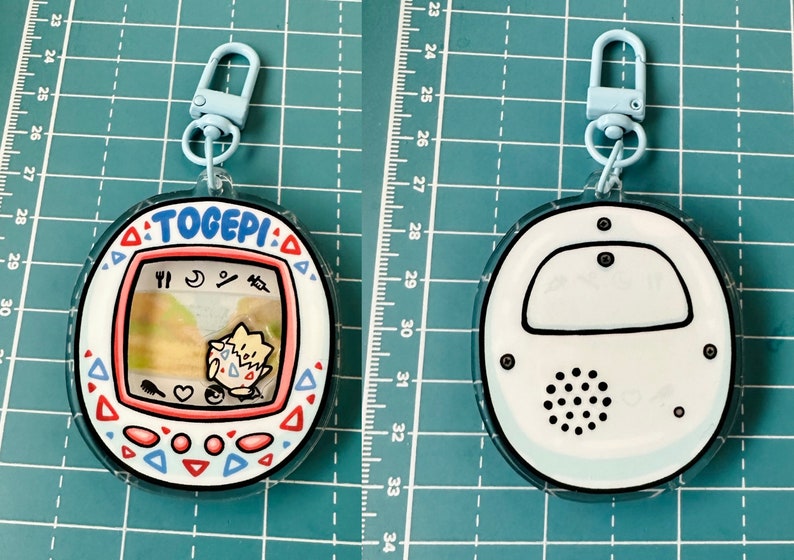May include: A white and blue plastic keychain with a Togepi character from the Pokemon franchise. The keychain is shaped like a Tamagotchi and has a small screen with a cartoon image of Togepi. The keychain has a blue clip and a black and white speaker on the back.