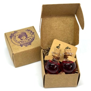 May include: Two small glass potion bottles filled with a red liquid, each with a cork stopper, presented in a small open cardboard box. A second, closed box with a purple illustration is visible. The bottles are displayed on a small brown tag.