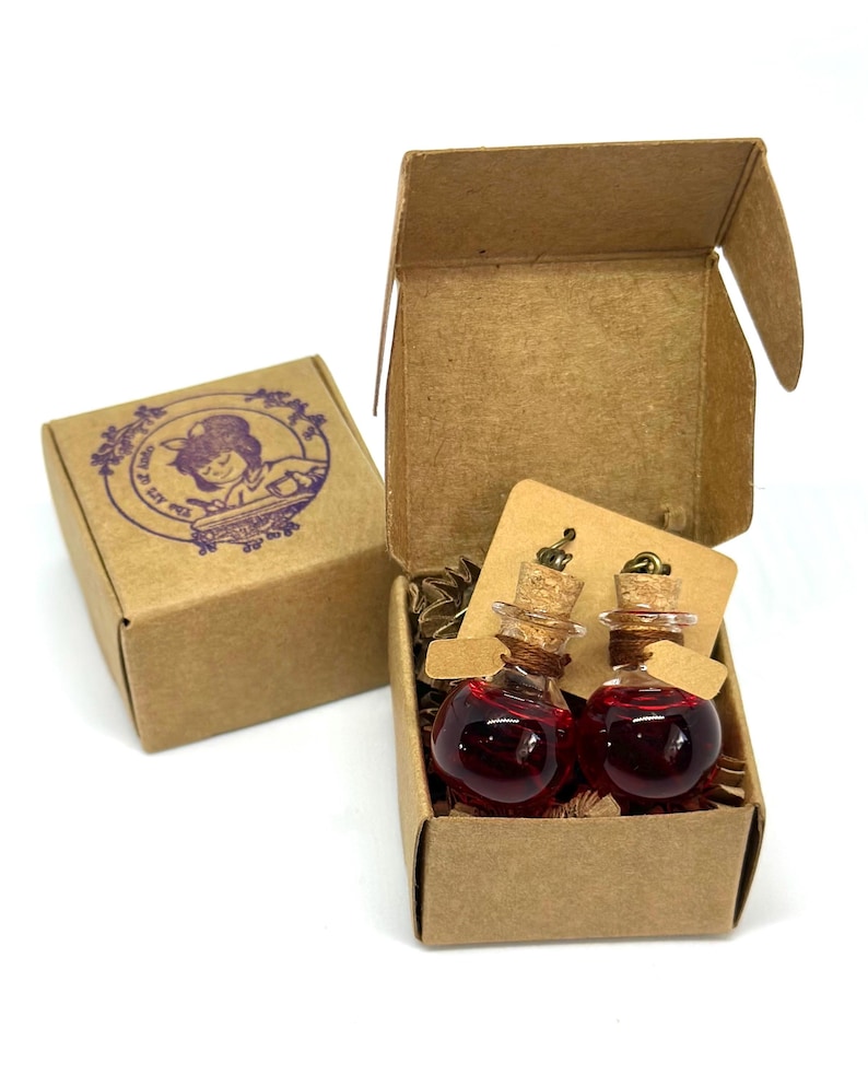 May include: Two small glass potion bottles filled with a red liquid, each with a cork stopper, presented in a small open cardboard box. A second, closed box with a purple illustration is visible. The bottles are displayed on a small brown tag.