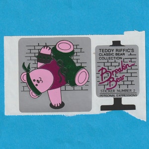 May include: A sticker featuring a pink bear wearing a bandana and a purple vest, with green accents. The sticker also includes the text "Teddy Riffic's Classic Bear Collection" and "Breakin' Bear" on a brick background. Sticker number 2.