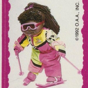 May include: A vintage Cabbage Patch Kids sticker showing a doll in a pink, yellow, and black ski suit, pink goggles, and skis. The doll has brown hair and is holding pink ski poles. The sticker is framed in a pink scalloped border.