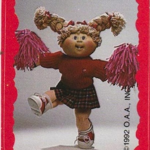 Op de afbeelding: Een vintage verzamelkaart met een Cabbage Patch Kid-pop verkleed als cheerleader. De pop draagt een rode trui, een geruite rok en witte sneakers. Ze houdt roze pompons vast en heeft gevlochten haar. De kaart heeft een rode rand en de tekst "©1992 O.A.A., INC."