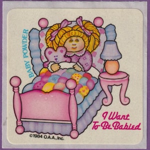 May include: A vintage sticker featuring a cartoon doll in a bed with a teddy bear. The doll has blonde hair and wears a purple nightgown. The bed is pink with a blue comforter. Text includes "Baby Powder" and "I Want To Be Babied."