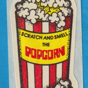 Puede incluir: Un cubo de palomitas de maíz a rayas rojas y blancas con palomitas de maíz amarillas en su interior. El cubo tiene el texto "Scratch and Smell the Popcorn" en la parte delantera.