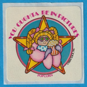 May include: A yellow star with a girl wearing pink sunglasses and a pink fur coat holding a red and white striped popcorn bucket. The text around the star reads "You Oughta Be In Pictures" and "Popcorn". The copyright text reads "©1984 O.A.A., Inc."