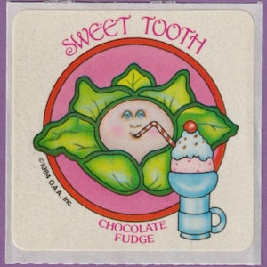 May include: A vintage sticker featuring a smiling face with a straw, surrounded by green leaves, and a sundae. The text reads "Sweet Tooth" and "Chocolate Fudge". The sticker has a pink and red circular design.