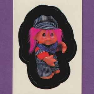 May include: A pink haired troll doll wearing a blue and white striped hat and overalls. The doll is standing on a black background.