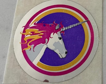 Puede incluir: Una pegatina circular con la cabeza de un unicornio de perfil. El unicornio tiene la cara blanca, la melena rosa y un cuerno en espiral. El diseño está sobre un fondo morado, enmarcado por anillos rosas, dorados y plateados. La pegatina está sobre un fondo cuadrado blanquecino.