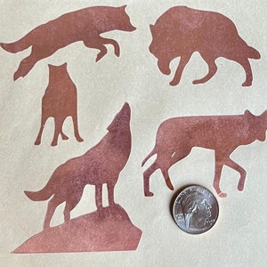 Puede incluir: Cinco siluetas de lobo de color cobre sobre un fondo beige. Los lobos están en varias poses, incluyendo aullar, correr y estar de pie. Una moneda de cuarto de dólar estadounidense de plata se incluye para la escala.