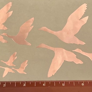May include: A set of nine copper-colored metal geese silhouettes in various sizes and positions. The geese are arranged in a cluster on a light brown background.