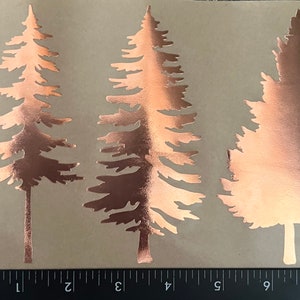 May include: Three copper-colored pine tree silhouettes on a brown background. The trees are cut out and have a metallic sheen.