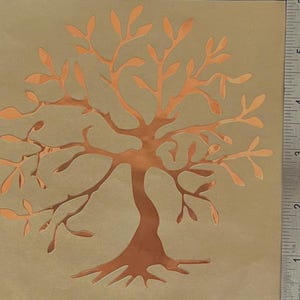 May include: A copper-colored tree stencil with a detailed design of branches and leaves. The stencil is on a beige background and measures approximately 5 inches tall.