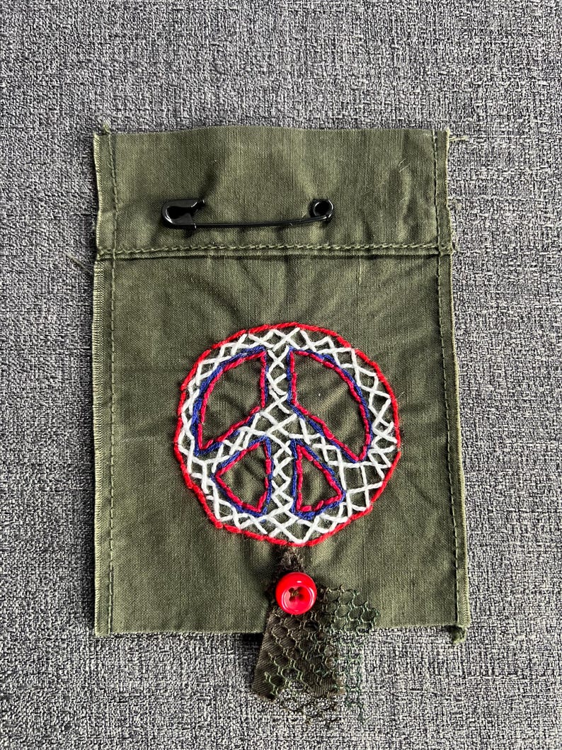 May include: Olive green fabric pouch with a hand-stitched peace sign in red, white, and blue thread. A black safety pin secures the top flap. A red button and a piece of green mesh are attached to the bottom.
