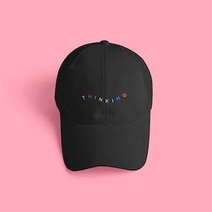 May include: Black baseball cap with the word "THINKING" embroidered in colorful letters across the front. The cap is set against a pink background.