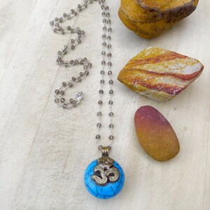 May include: A long necklace with a silver chain and small gray beads. The pendant is a round, turquoise-colored stone with a silver-colored Om symbol. The necklace is displayed on a light wooden surface with decorative stones.