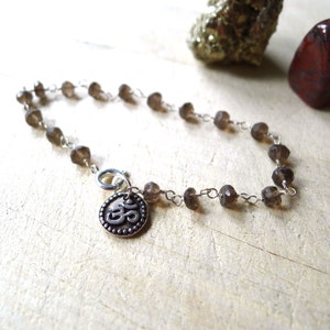 May include: A brown gemstone bracelet with a silver Om charm. The bracelet is made with small, faceted gemstones and a silver chain.