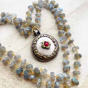May include: A handcrafted necklace featuring a pendant with a silver center and a red gemstone flower. The pendant is surrounded by an ornate gold-colored frame. The necklace is made of small, gray-blue gemstone beads strung on gold-colored wire.