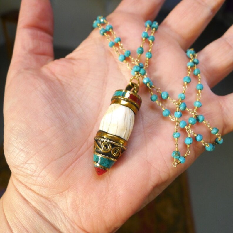 K&ouml;nnte beinhalten: Eine goldene Kettenkette mit einer Kette aus t&uuml;rkisfarbenen Perlen und einem Anh&auml;nger mit einem wei&szlig;en knochenartigen Zylinder mit goldenen und t&uuml;rkisfarbenen Akzenten.