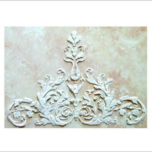 May include: White, ornate, raised relief design featuring a symmetrical arrangement of stylized leaves and scrolls. The design is centered on a textured, light-colored background, creating a classic, decorative element.