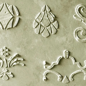 May include: Five white, raised decorative elements on a muted green background. The designs include a diamond shape, a teardrop shape, a scroll, a floral design, and a symmetrical design with curved lines and a triangle.