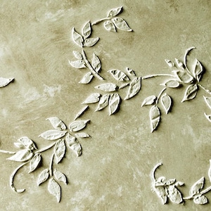 May include: Close-up of a textured, neutral-toned surface with white, raised floral designs. The design features stylized leaves and vines arranged in a circular and linear pattern, creating a decorative, organic aesthetic.
