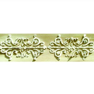 May include: Two ornate, white decorative architectural elements with scrolling floral designs. The elements are symmetrical and appear to be made of a carved material, possibly wood or plaster. They are set against a light beige background.