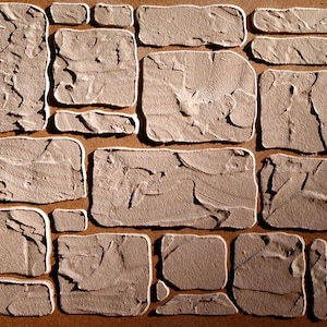 May include: A close-up view of a textured, brown, and beige stone wall pattern. The stones are rectangular and square, with a rough, uneven surface. The grout lines are white, creating a contrast with the stone.