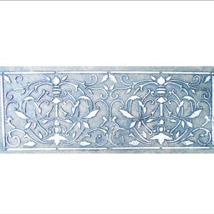 May include: A rectangular, light blue decorative panel with an ornate, symmetrical design. The design features scrolling vines, leaves, and stylized floral elements in white, creating a textured, embossed appearance. The panel has a border.
