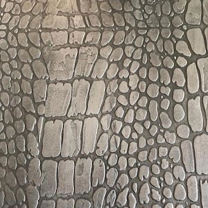 May include: Close-up of a textured, metallic surface with a pattern resembling crocodile skin. The design features irregular, rectangular and oval shapes in varying sizes, creating a rugged, industrial aesthetic. The color is a muted silver-gray.