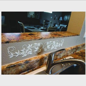 May include: A kitchen countertop with a decorative white leaf and vine design. The countertop has a brown and tan speckled pattern. A stainless steel faucet is in the foreground.