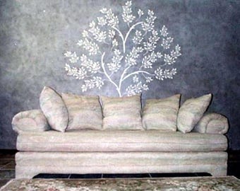 May include: A beige sofa with rolled arms and multiple throw pillows. The sofa is positioned in front of a textured gray wall with a white tree design. A patterned rug is visible in the foreground.