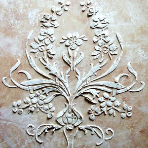 May include: A detailed, raised relief floral design in white against a mottled, tan background. The symmetrical composition features stylized flowers, leaves, and curved elements, creating an elegant, textured pattern. The design is reminiscent of classic decorative art.