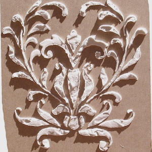 May include: A detailed, raised white floral design against a tan background. The design features stylized leaves and swirling patterns, creating a textured, three-dimensional effect. The artwork is centered and symmetrical.