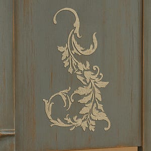 May include: Close-up of a section of a blue-gray wooden cabinet. A decorative, gold-colored floral design is stenciled on the cabinet door. The cabinet has a distressed finish, with visible wood grain.