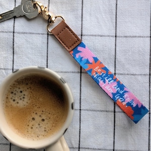 Könnte beinhalten: Ein Schlüsselband mit einem Blumenmuster in Blau, Rosa und Orange, mit dem Text "Best is yet to come". Das Schlüsselband hat einen braunen Lederakzent und einen goldfarbenen Verschluss. Eine Tasse Kaffee steht neben dem Schlüsselband.
