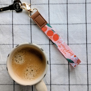 以下が含まれることがあります： ピンク色のキーリストレット。花柄とオレンジ色のデザインで、ゴールドカラーの留め金と茶色のレザータブが付いています。リストレットは、コーヒーカップと鍵の隣にあり、白と黒のチェック柄の表面に置かれています。