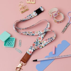 Op de afbeelding: Een witte lanyard met een bloemenpatroon en de tekst "All things are possible with God" in bruine letters. De lanyard heeft een bruin leren bandje en een gouden metalen clip.