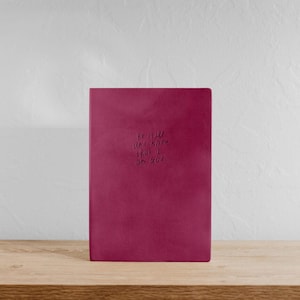 May include: A burgundy journal with the embossed text "Be still and know that I am God." The journal is standing upright on a wooden shelf against a white wall. The cover is a solid, smooth colour.