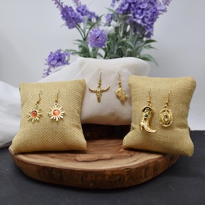 May include: A collection of gold-colored earrings displayed on small burlap pillows. The earrings feature various designs, including a sunburst, a bull skull, a cactus, a cowboy boot, and an oval shape. The earrings are presented on a wooden tray.