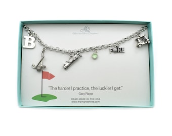 May include: A silver charm bracelet with golf-themed charms, including a golf club, golf bag, golf cart, and the letter "B". The bracelet is displayed in a light blue box with a quote by Gary Player.